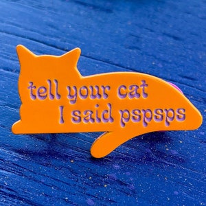 Puede incluir: Pin de metal naranja con forma de gato con el texto "tell your cat I said pspspps" en morado.