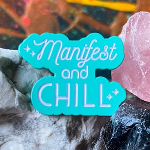 May include: A turquoise blue enamel pin with the text "Manifest and Chill" in white lettering. The pin is surrounded by small white stars and is sitting on top of a white rock.