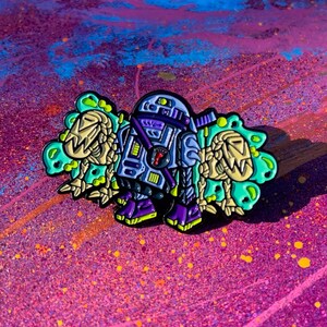 Puede incluir: Un robot morado y azul con una criatura alienígena amarilla y verde a cada lado. El robot tiene una cabeza morada con un ojo negro y una antena negra. El robot lleva un cinturón negro con una hebilla morada. El robot está de pie sobre un fondo rosa y azul.