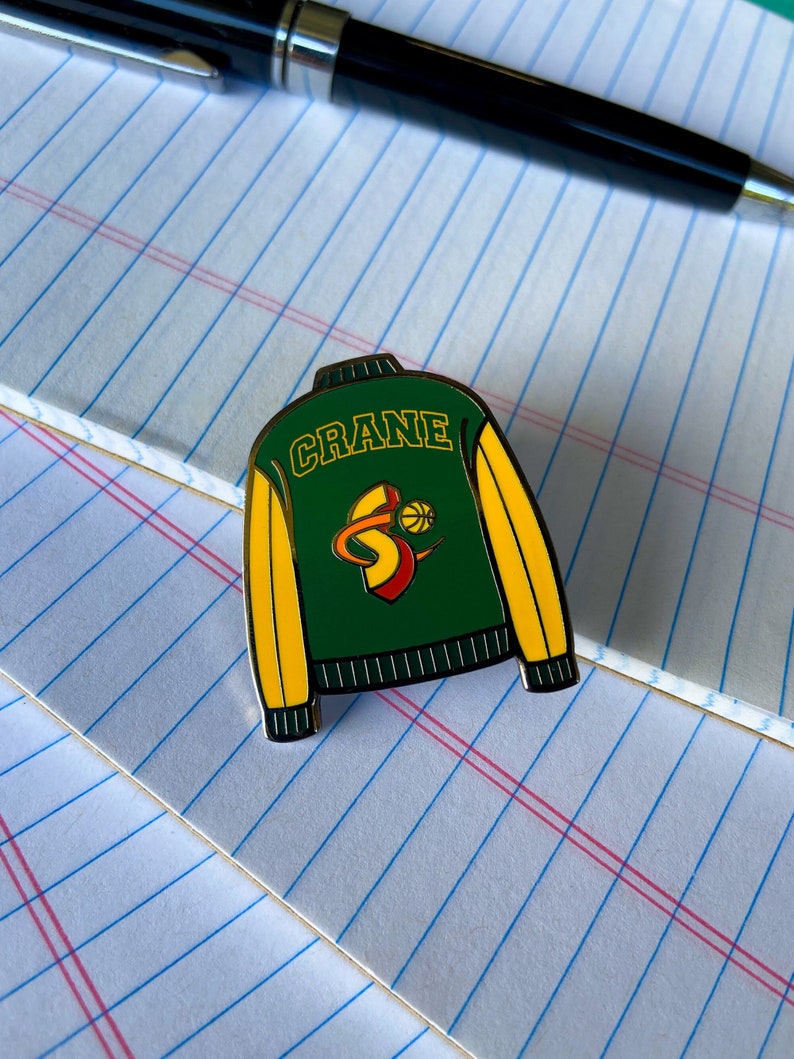 May include: A green and yellow enamel pin shaped like a letterman jacket with the word "CRANE" and a basketball logo on the front.
