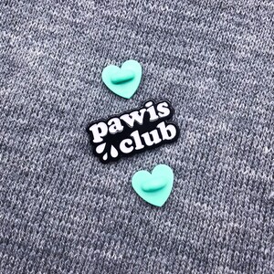 Peut inclure: Deux &eacute;pingles en forme de c&oelig;ur turquoise avec une &eacute;pingle noire avec du texte blanc qui dit "paw is club" sur un fond textur&eacute; gris.