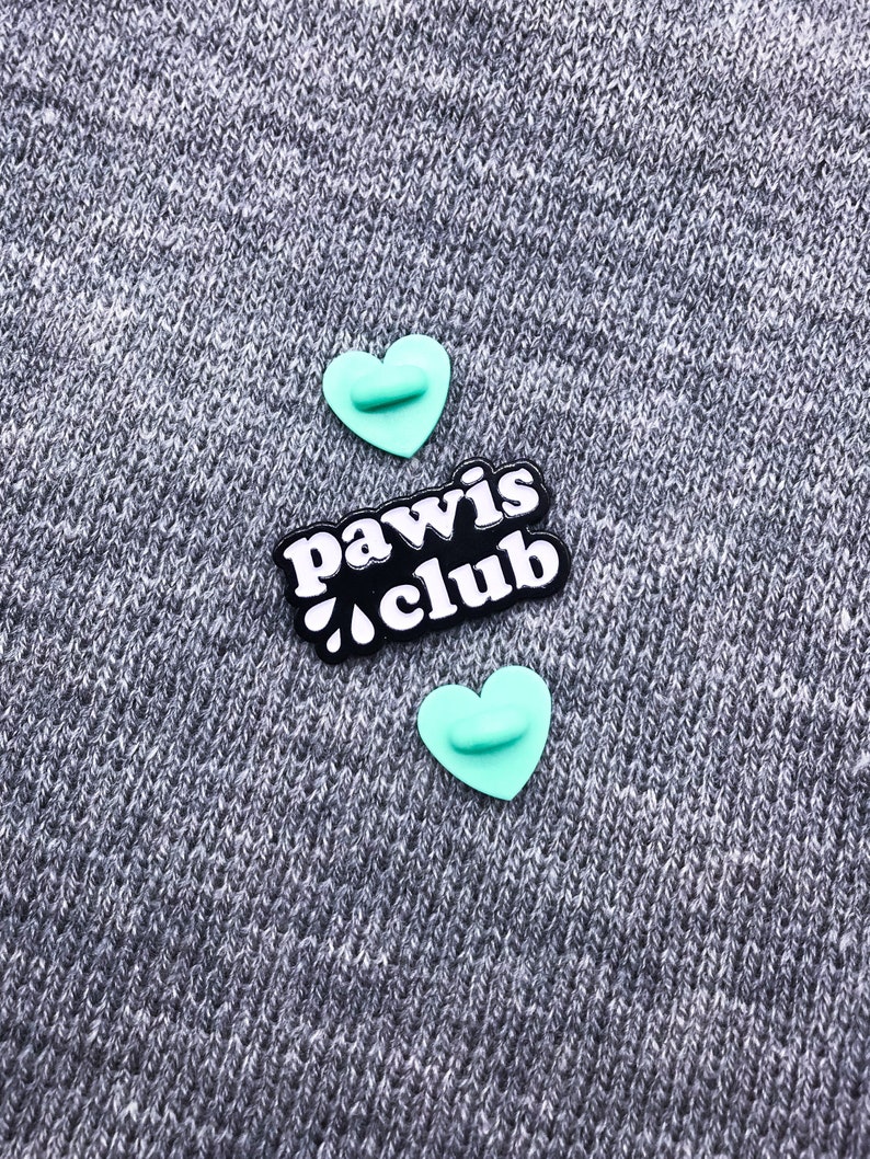 Peut inclure: Deux &eacute;pingles en forme de c&oelig;ur turquoise avec une &eacute;pingle noire avec du texte blanc qui dit "paw is club" sur un fond textur&eacute; gris.