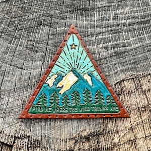 May include: A triangular leather patch with a mountain scene and a star. The patch is brown with a blue, green, and white mountain scene. The text "Find me where the wild things are" is printed on the bottom of the patch.
