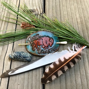 May include: An oval, turquoise leather hair barrette with a carved bison and the words "Roam Wild & Free" in white. The barrette is next to a pine branch, feathers, and a stone on a wooden surface.
