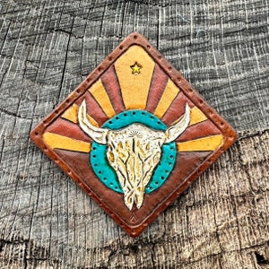 May include: A leather patch with a diamond shape features a white bull skull with a turquoise background. The skull is surrounded by a sunburst design with red and yellow rays.