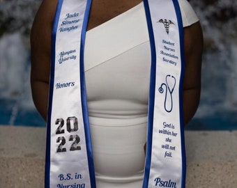 May include: White graduation sash with blue trim, featuring the text "Jada Simone Vaughn", "Hampton University", "Honors", "2022", "B.S. in Nursing", "Student Nurses Association Secretary", "God is within her she will not fail", and "Psalm 46:5".
