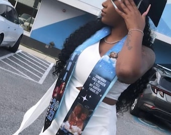 May include: A woman wearing a white dress and a blue and black graduation sash with a photo of a woman and a child. The sash has the text "Somebody call heaven & tell my aunties I made it!" and "Rest in Love Auntie".