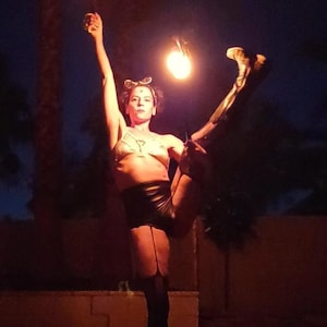 May include: A person performing with fire at night. The person is wearing a bra, shorts, and stockings, and is holding two flaming torches. The background is dark with a few trees and a building.