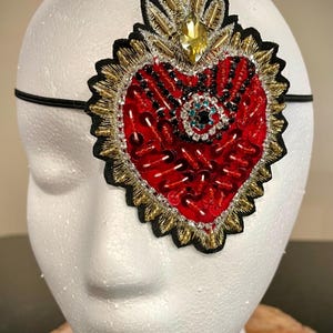 May include: A red beaded heart with gold and black embroidery and a large jewel in the center. The heart is attached to a black elastic headband.