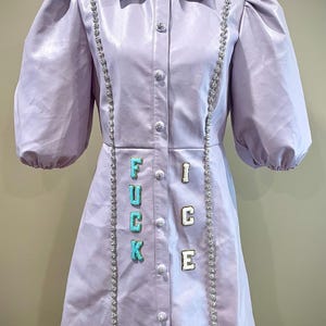 May include: A lavender-colored dress with a collared neckline and button-down front. The dress features puff sleeves, decorative rhinestone trim, and the word "FUCK ICE" spelled out in colorful letters. A heart-shaped patch is near the hem.