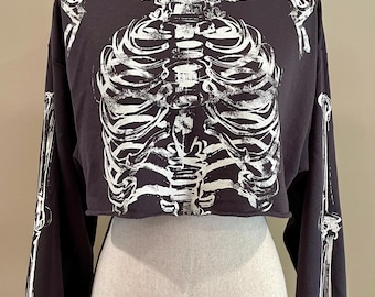 May include: Dark gray, long-sleeved crop top featuring a white skeleton design on the front and arms. The top has a high neckline and a cropped hemline, perfect for Halloween or gothic-themed outfits.