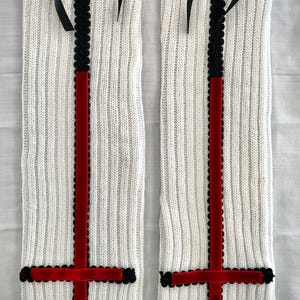 May include: Pair of white knit leg warmers with black ribbon bows at the top. Each leg warmer features a vertical black lace trim and a red velvet cross design. The leg warmers are displayed on a light background.
