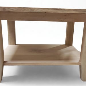 Könnte beinhalten: Ein helles Couchtisch aus Holz mit einem unteren Regal. Der Tisch hat eine rechteckige Oberseite und vier stabile Beine. Die Beine sind leicht nach außen abgewinkelt und bieten Stabilität. Der Tisch ist aus unbehandeltem Holz gefertigt.