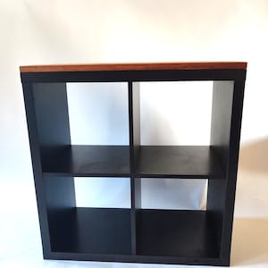 Könnte beinhalten: Ein schwarzes, würfelförmiges Aufbewahrungsregal mit vier Fächern. Die Oberseite des Regals ist warmbraun und kontrastiert mit dem schwarzen Rahmen. Das Regal dient zum Ordnen und Präsentieren von Gegenständen.