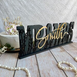 May include: Black wooden wedding cake topper with gold lettering that reads "Mr. & Mrs. Smith" with floral accents.