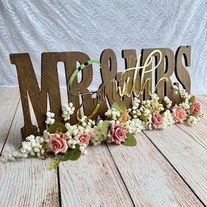 May include: Wooden "Mr & Mrs Smith" wedding sign with gold lettering and a floral arrangement of white and pink flowers.