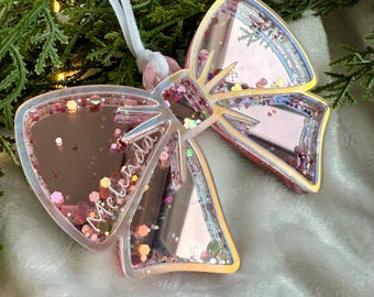May include: A pink and iridescent glitter bow ornament with the name "Melinda" inscribed on it.