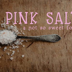 May include: Pink Himalayan salt in a silver spoon on a wooden surface. The salt is scattered around the spoon. The text "PINK SALT a not so sweet font" is written in pink letters above the spoon.