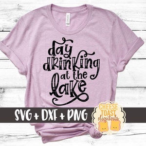 May include: A pink t-shirt with the text "day drinking at the lake" in black. The shirt is on a white wood background. The text "SVG + DXF + PNG" is below the design. The text "CHEESE TOAST digitals" is in a yellow circle with a black outline.