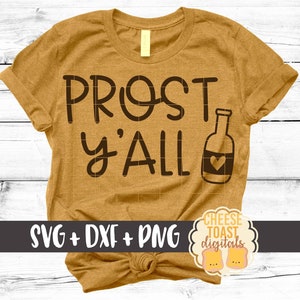 以下が含まれることがあります： マスタードイエローのTシャツに「Prost y'all」の文字と、ハートの付いたビール瓶のシルエットがプリントされています。Tシャツは裾が結ばれています。