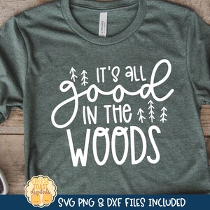 Può includere: Una maglietta verde con testo bianco che recita "It's all good in the woods" con un disegno di piccoli alberi. La maglietta è su una superficie di legno con uno zaino e una coperta di lana.
