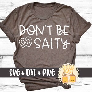 以下が含まれることがあります： プレッツェルのハートのグラフィックが入った、白い文字で「Don't be Salty」と書かれた茶色のTシャツ。Tシャツの下部に「SVG + DXF + PNG」の文字があります。