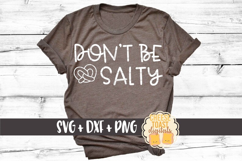 以下が含まれることがあります： プレッツェルのハートのグラフィックが入った、白い文字で「Don't be Salty」と書かれた茶色のTシャツ。Tシャツの下部に「SVG + DXF + PNG」の文字があります。