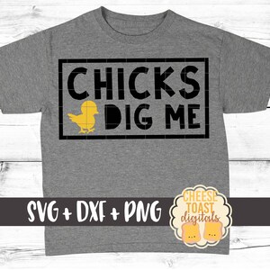 May include: A grey t-shirt with black text that reads "CHICKS DIG ME" and a yellow chick graphic. The text is in a box with a black border. The t-shirt is on a white wood background.