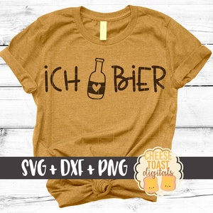 以下が含まれることがあります： 黒文字で「ICH &hearts; BIER」と書かれたマスタードイエローのTシャツ。Tシャツは裾が結ばれており、白い木製の背景で撮影されています。
