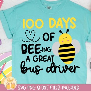 Op de afbeelding: Een lichtblauw t-shirt met de tekst "100 DAYS OF BEE-ING A GREAT bus driver" in geel en zwart. Een cartoon bijenafbeelding staat op de shirt. Schoolspullen, waaronder een liniaal, schaar en potloden, staan op de achtergrond.