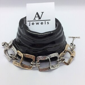 May include: A silver and gold tone bracelet with square links. The bracelet is displayed on a black ceramic stand with a white card that reads "A jewels".