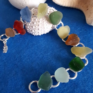 Könnte beinhalten: Ein silbernes Kettenarmband mit bunten Meerglassperlen. Die Perlen sind in verschiedenen Grüntönen, Blau, Rot und Gelb. Das Armband liegt auf einem blauen Hintergrund mit einem weißen Seeigel und einem Seestern.