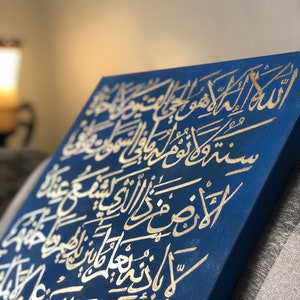 May include: A blue canvas with gold Arabic calligraphy. The calligraphy is in a traditional style and is arranged in a vertical format. The canvas is a square shape and has a textured surface.