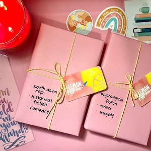 Puede incluir: Dos libros envueltos en papel rosa y cuerda amarilla. Cada libro tiene una etiqueta en forma de limón con el texto "Lemon Pop Shop" y una descripción del género del libro. El primer libro se describe como "Representación del sur de Asia, ficción histórica, romance". El segundo libro se describe como "Ficción psicológica, thriller, tragedia."