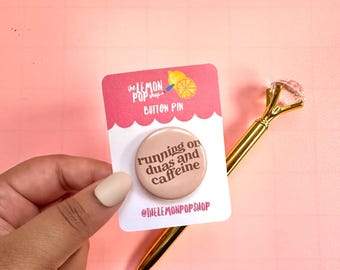 Könnte beinhalten: Ein Button-Pin von The Lemon Pop Shop mit dem Text "running on duas and caffeine" in Braun auf hellbraunem Hintergrund. Der Pin ist an einer rosa-weißen Karte mit dem Logo des Shops befestigt. Ein goldener Stift mit diamantförmigem Aufsatz befindet sich im Hintergrund.