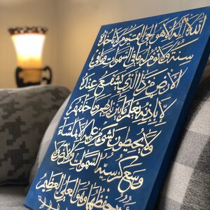 May include: A blue canvas with gold Arabic calligraphy. The calligraphy is on a dark blue background and is framed with a simple gold border. The canvas is leaning against a gray and white patterned couch.