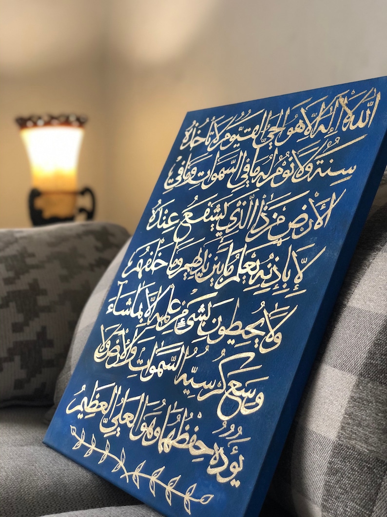 May include: A blue canvas with gold Arabic calligraphy. The calligraphy is on a dark blue background and is framed with a simple gold border. The canvas is leaning against a gray and white patterned couch.