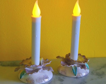 May include: Two white candle holders with a floral design. Each holder has a white candle with a yellow flame.