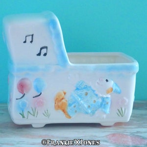 May include: A white ceramic planter shaped like a baby crib with blue and pink accents. The planter features a musical note design on the headboard and a painted image of a baby in a blue and white onesie with a balloon.