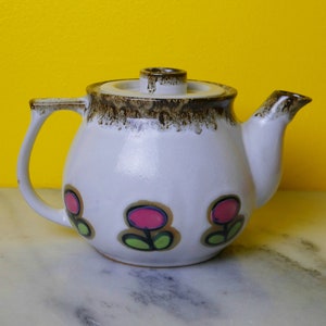 Peut inclure: Une théière en céramique blanche avec un couvercle et une bordure marrons. La théière est décorée d'un motif floral avec des fleurs roses et vertes.