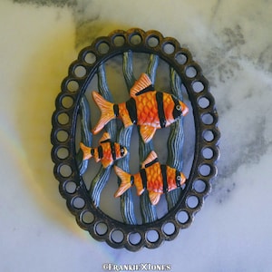 May include: A round, metal, decorative trivet with a scalloped edge. The trivet features three orange and black fish swimming among blue and white seaweed.