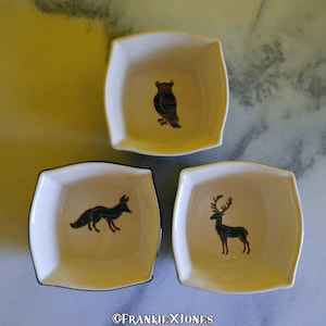 May include: Three small white ceramic square dishes with black rims. Each dish features a gold silhouette of an animal: an owl, a fox, and a deer.
