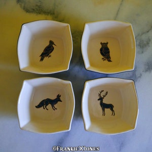 Pode incluir: Conjunto de quatro pratos quadrados de cerâmica branca com acabamento preto. Cada prato apresenta uma silhueta preta de um animal diferente: um pica-pau, uma coruja, uma raposa e um veado.