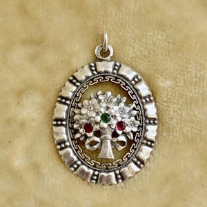 Pode incluir: Pingente de prata com design floral. O pingente apresenta um buquê de flores com uma gema verde e duas vermelhas. Tem formato oval com borda decorativa e laço para corrente.