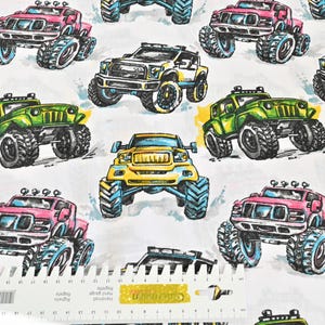 Peut inclure: Tissu blanc avec un motif répété de monster trucks colorés. Les camions sont dans des tons de rose, jaune et vert, avec de gros pneus et des motifs détaillés. Le fond est de couleur claire et neutre.