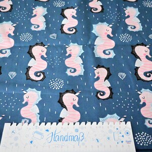 Peut inclure: Tissu bleu avec un motif répété de hippocampes roses avec des contours noirs et des accents blancs. Le tissu a une règle à motifs blancs, noirs et bleus sur le dessus avec le mot "Handmade" écrit dessus.
