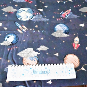 Könnte beinhalten: Ein marineblauer Stoff mit einem sich wiederholenden Muster aus Planeten, Sternen, Raketen und Wolken. Die Planeten sind in Blautönen, Braun und Weiß gehalten. Die Raketen sind rot und weiß. Die Wolken sind weiß. Die Sterne sind golden. Der Stoff hat ein "Handmade"-Etikett.