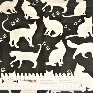 Könnte beinhalten: Schwarz-weißes Stoff mit einem sich wiederholenden Muster aus weißen Katzen-Silhouetten und Pfotenabdrücken. Ein Lineal mit dem Text "Gütermann Creativ" befindet sich am unteren Rand des Bildes.