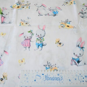 Könnte beinhalten: Weißer Stoff mit einem sich wiederholenden Muster aus Hasen und Blumen. Die Hasen sind weiß mit rosa und blauen Akzenten. Die Blumen sind gelb und blau. Der Stoff hat den Text "Handmade" aufgedruckt.