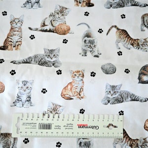 Könnte beinhalten: Weißer Stoff mit einem sich wiederholenden Muster von Kätzchen, die mit Garn spielen. Die Kätzchen sind in verschiedenen Posen und Farben, darunter Tabby, Calico und Grau. Der Stoff ist mit schwarzen Pfotenabdrücken verziert.